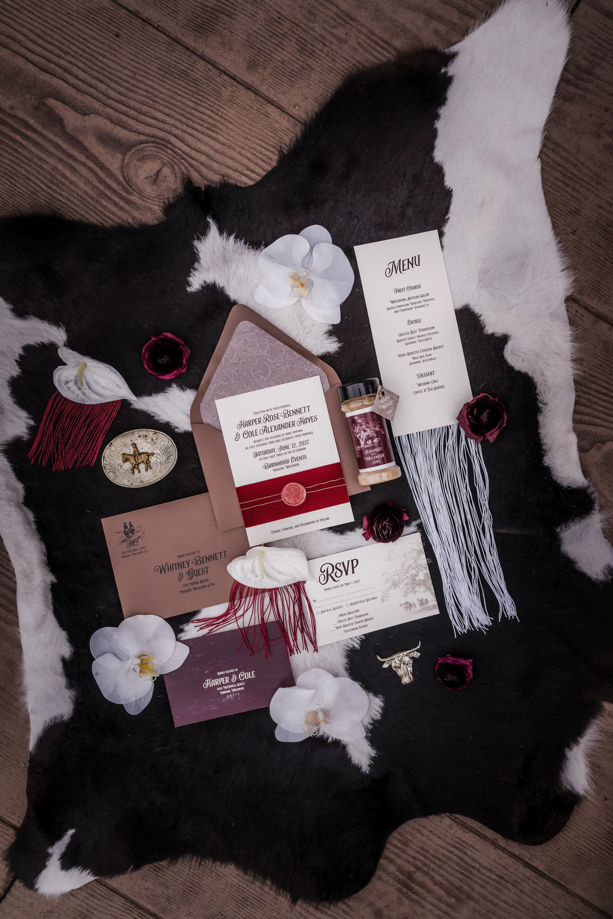 Western cowboy-themed wedding stationery and printed pieces from LK Bridal Studio in cream, taupe, and burgundy. Included pieces are an invitation with velvet belly band with gold thread and wax seal, RSVP with horse illustration, printed envelope and envelope liner, and tasseled dinner menu. The pieces are photographed on a cowhide rug alongside belt buckle, florals, and custom spice favor with hang tag