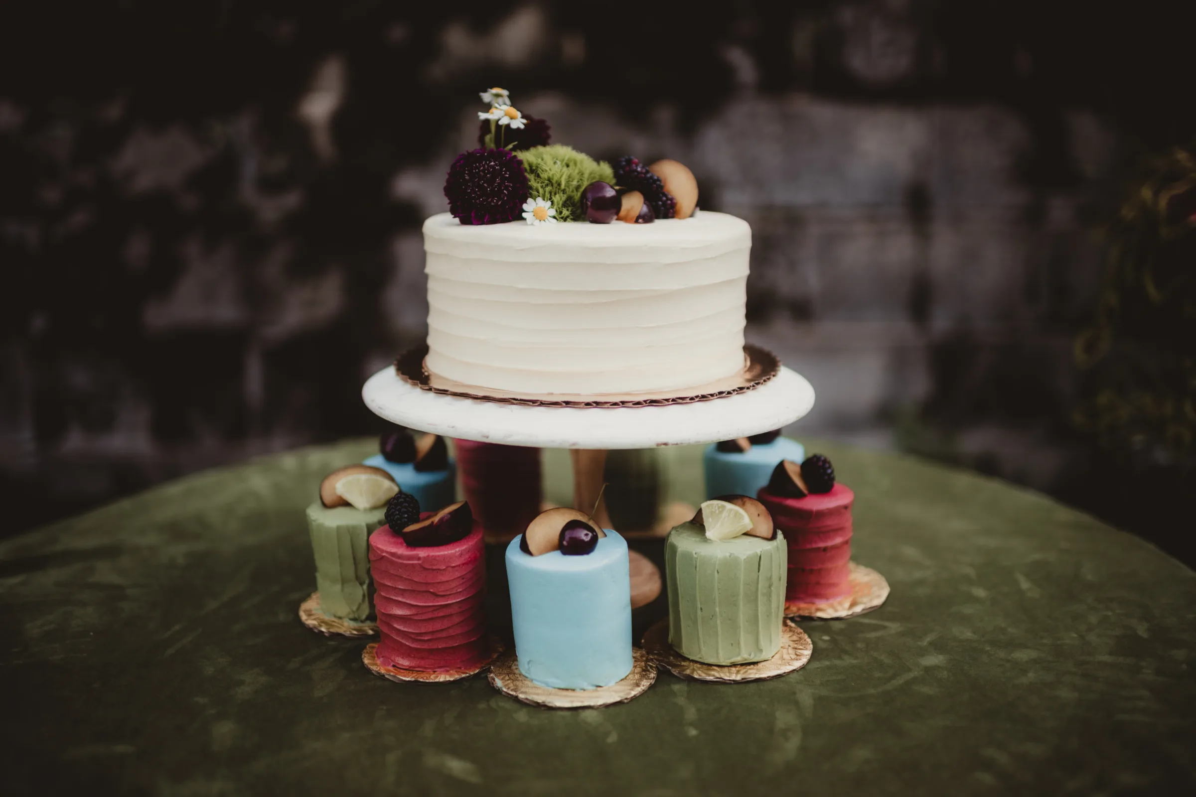 cake and dessert inspiration from Sandy Kakes showing a single tier white wedding cake on a pedestal surrounded by textured, colorful, single-serving cakes. All are topped with fruit and/or florals