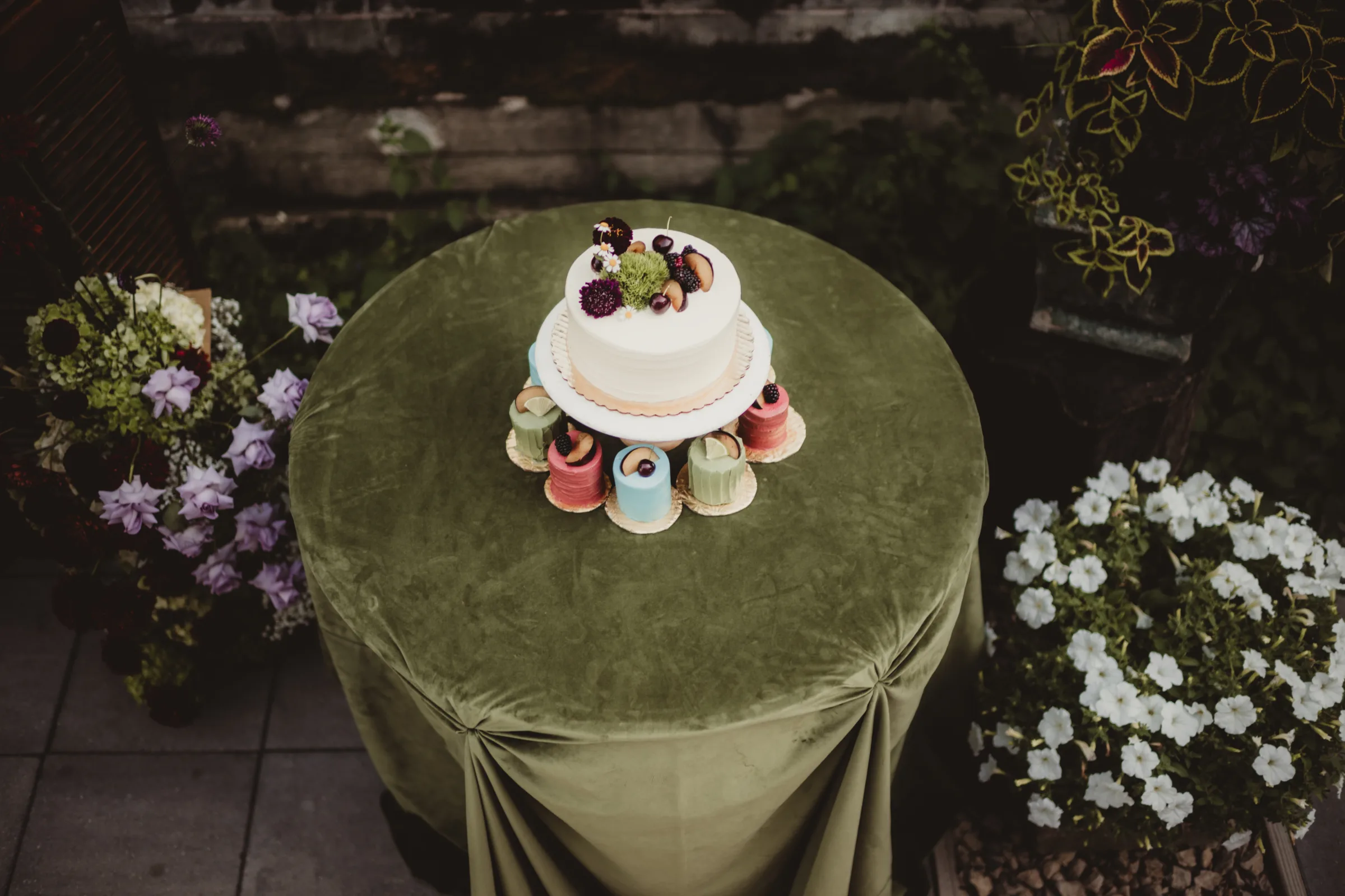 cake and dessert inspiration from Sandy Kakes showing a single tier white wedding cake on a pedestal surrounded by textured, colorful, single-serving cakes. All are topped with fruit and/or florals