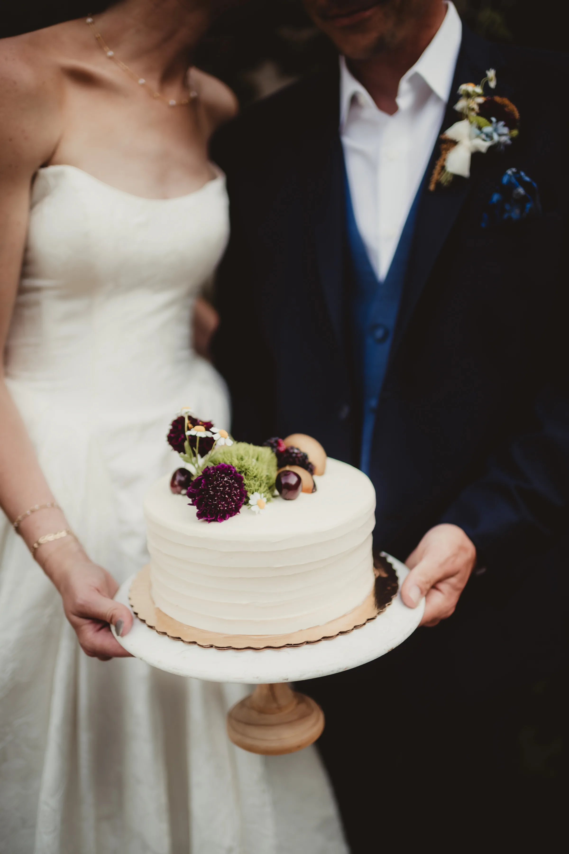 the bride and groom models present a single tier white wedding cake from Sandy Kakes topped with fruit and florals