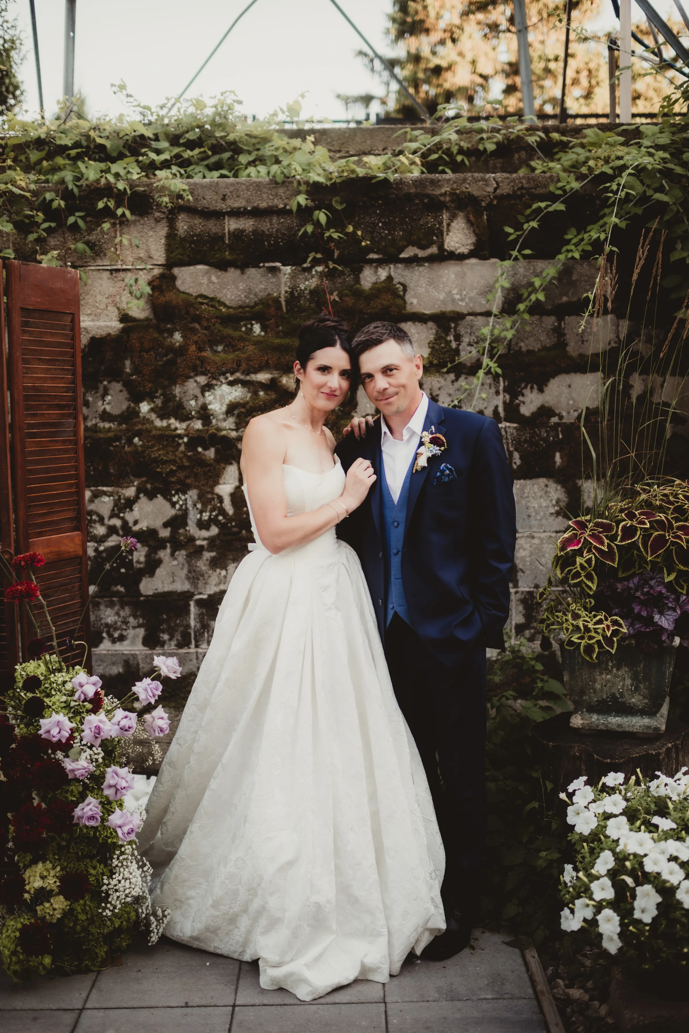 the couple poses in front of a brick wall covered in greenery and surrounded by florals