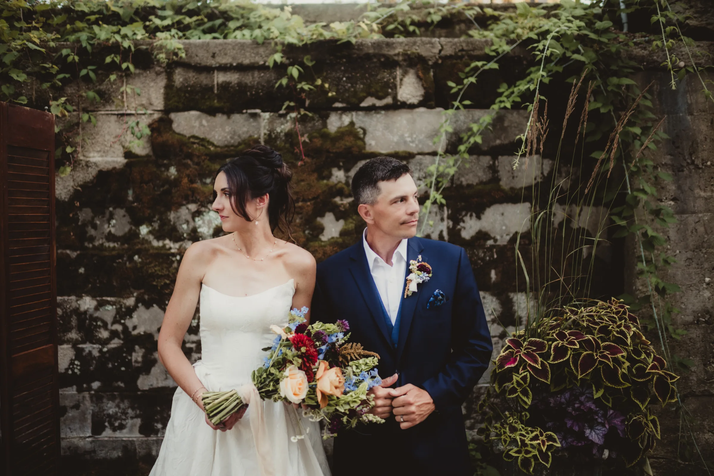 the couple poses in front of a brick wall covered in greenery, with the bride holding a bouquet from Signature Events