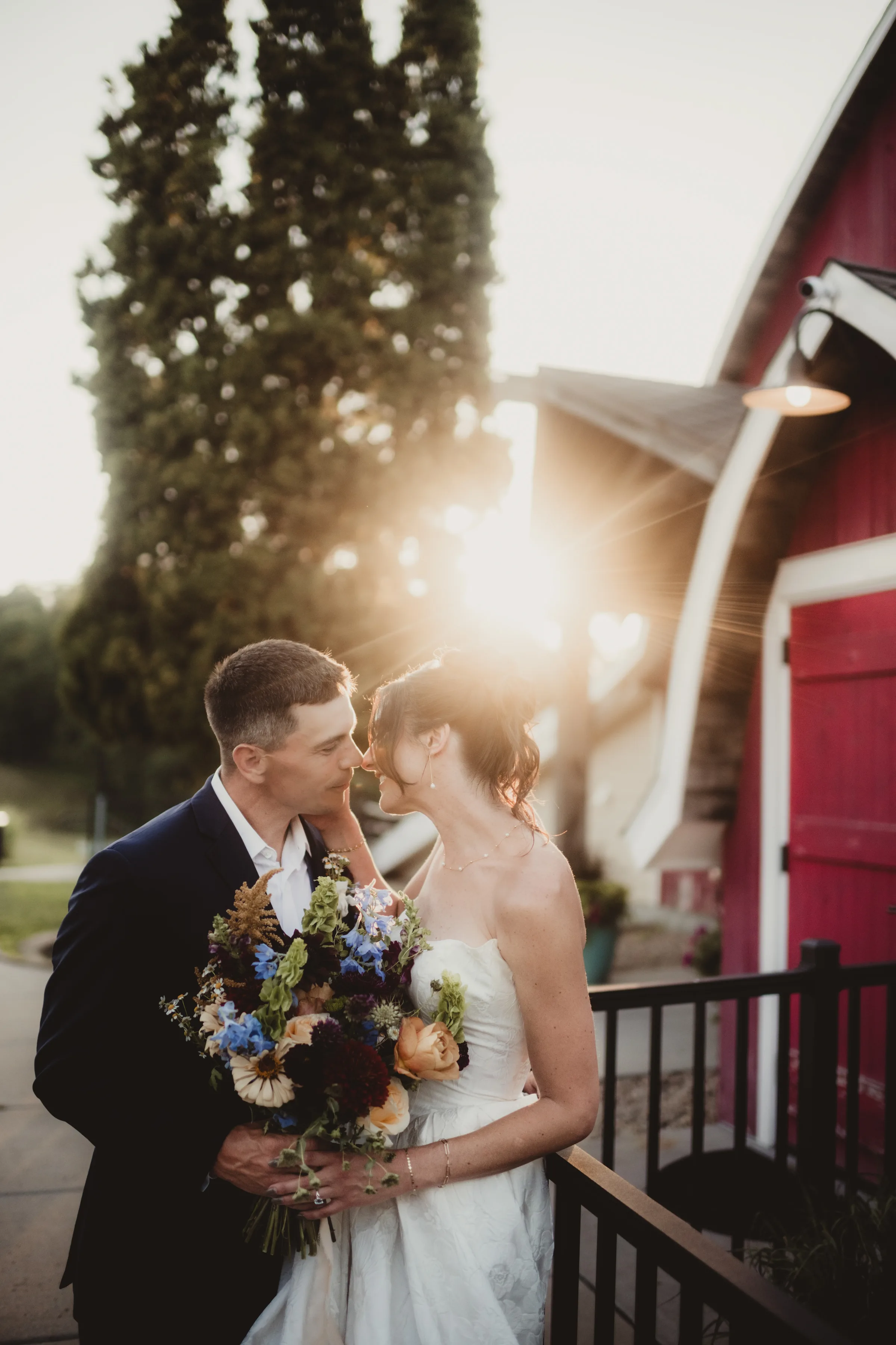 a couple portrait at golden hour, with sun peeking through the trees beside the red barn at Vennebu Hill