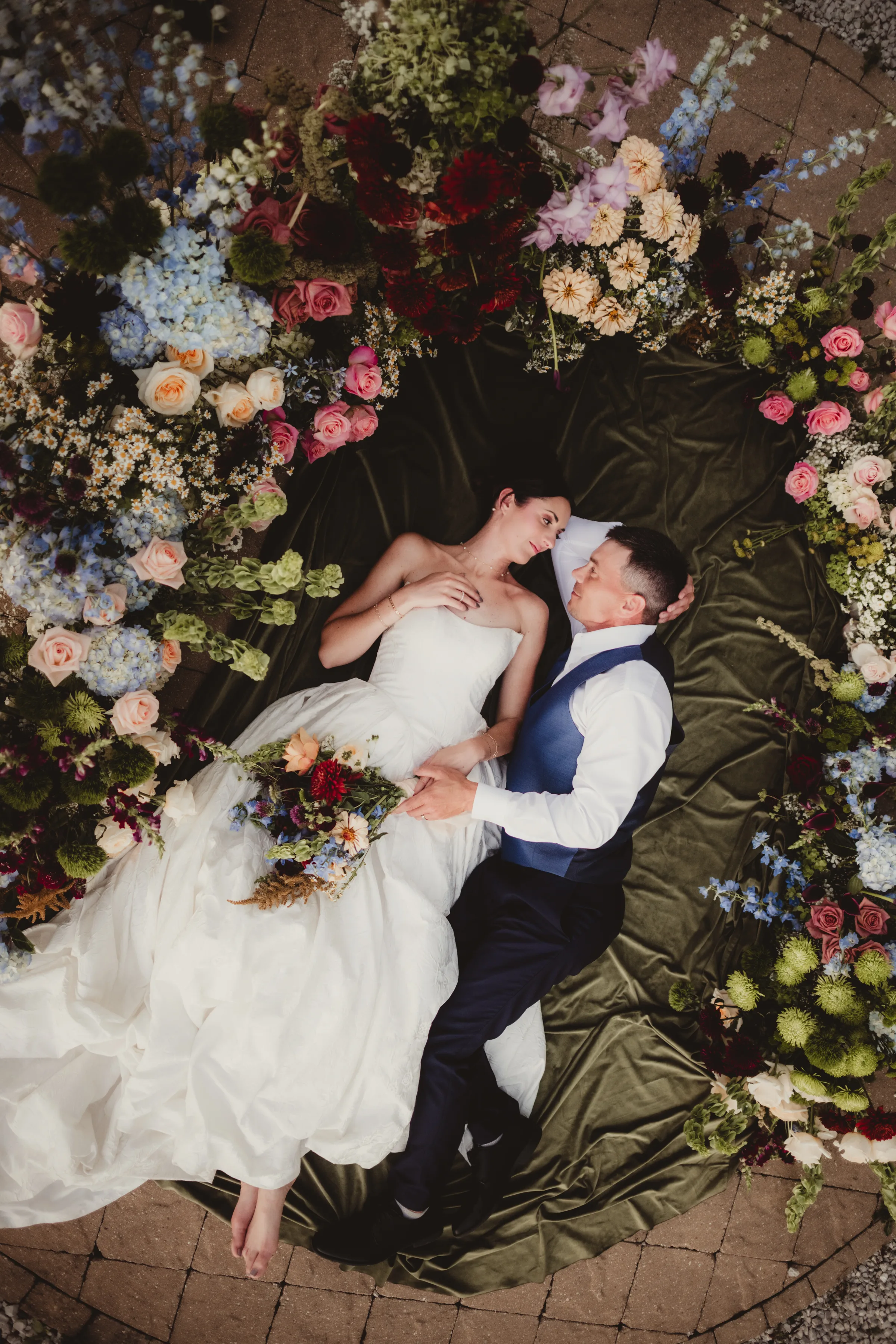 an artful, overhead photo of the bride and groom models lying on a green blanket surrounded by an arch of spiced garden party wildflower inspired florals from Signature Events