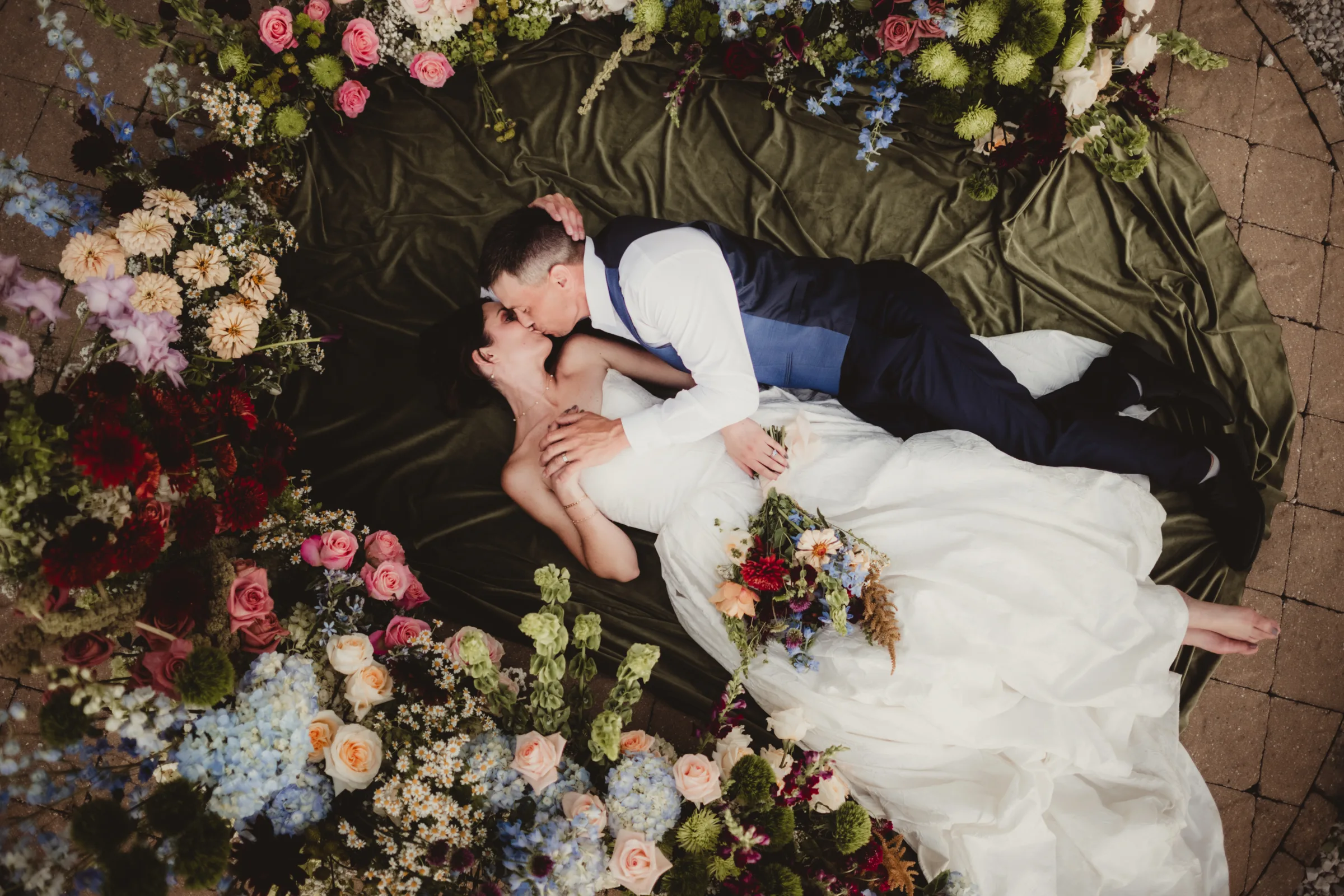 an artful, overhead photo of the bride and groom models lying on a green blanket surrounded by an arch of wildflower inspired florals from Signature Events