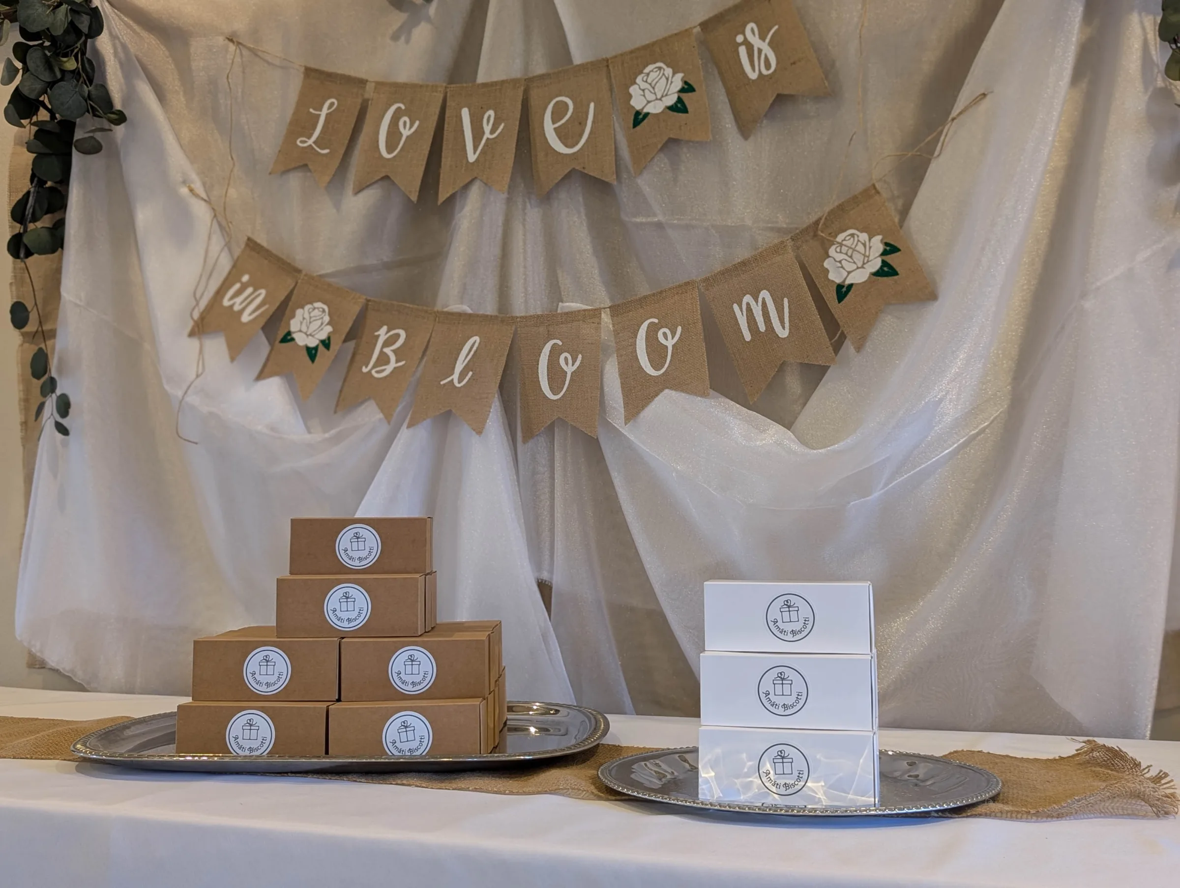 Amati Biscotti favor boxes stacked on silver trays in front of a "Love is in Bloom" burlap banner at a bridal shower