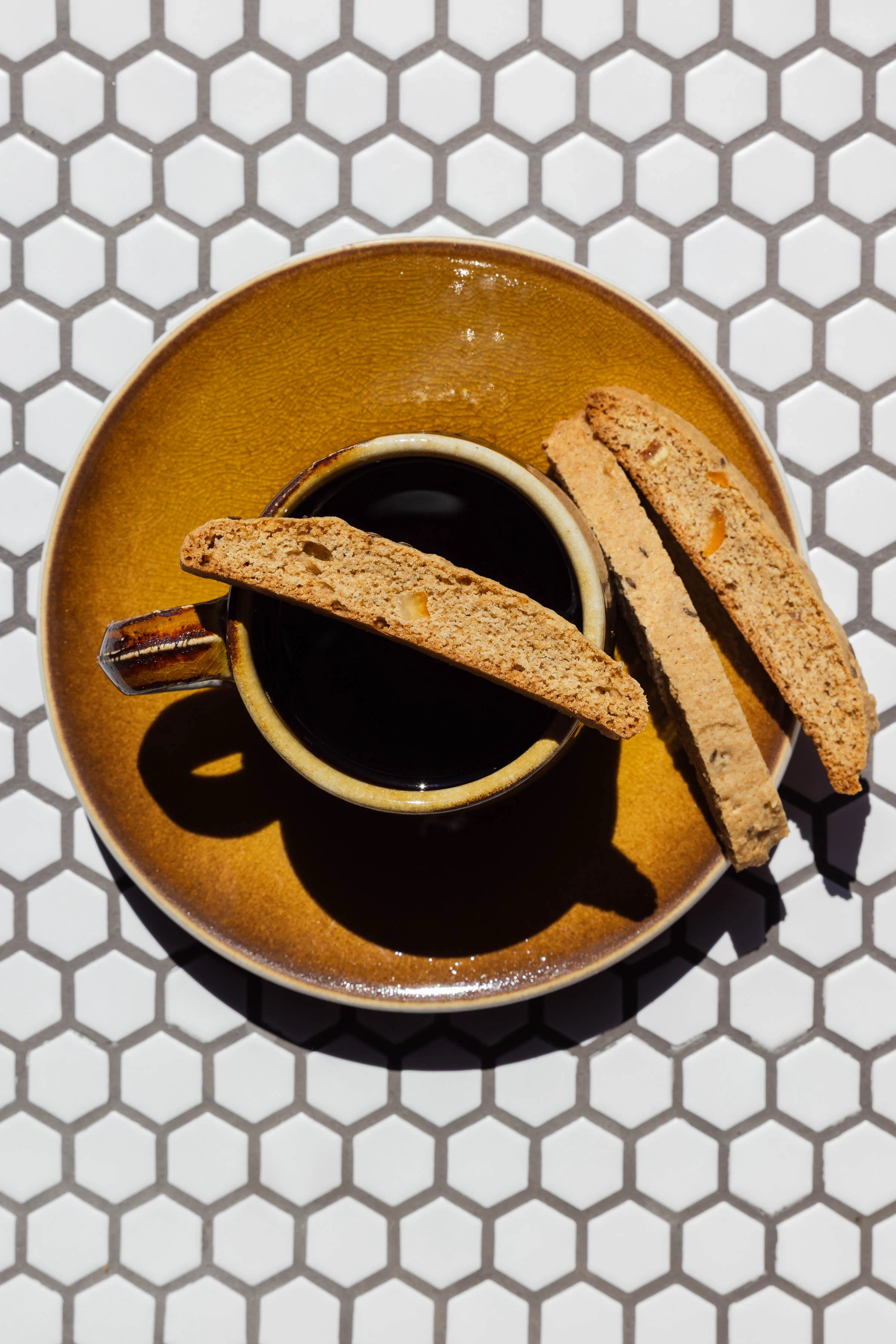 Amati Biscotti biscotti served alongside a cup of coffee on a hexagon tile background