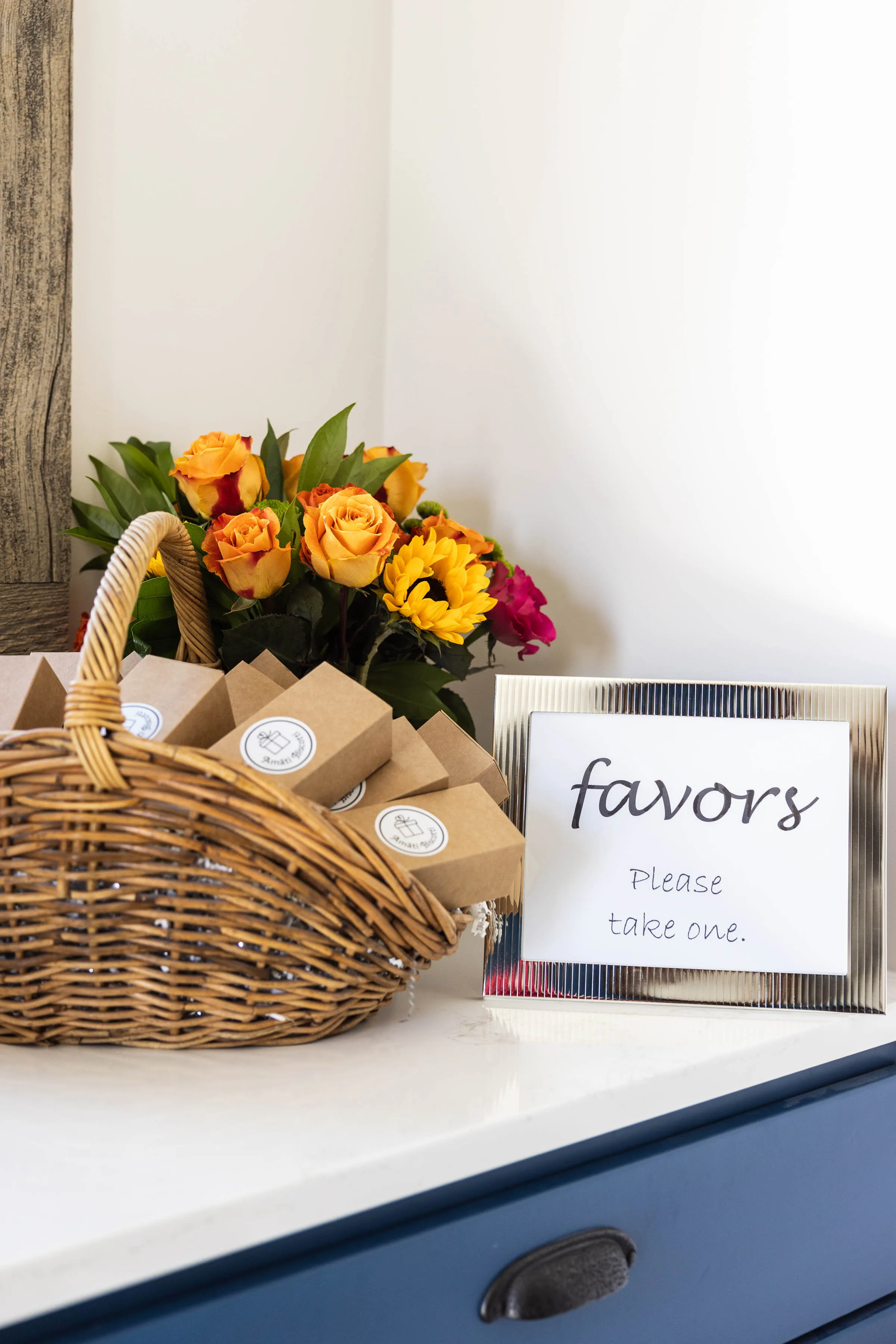 Amati Biscotti wedding favor boxes in a basket with sunflowers and roses beside a "favors, please take one" sign