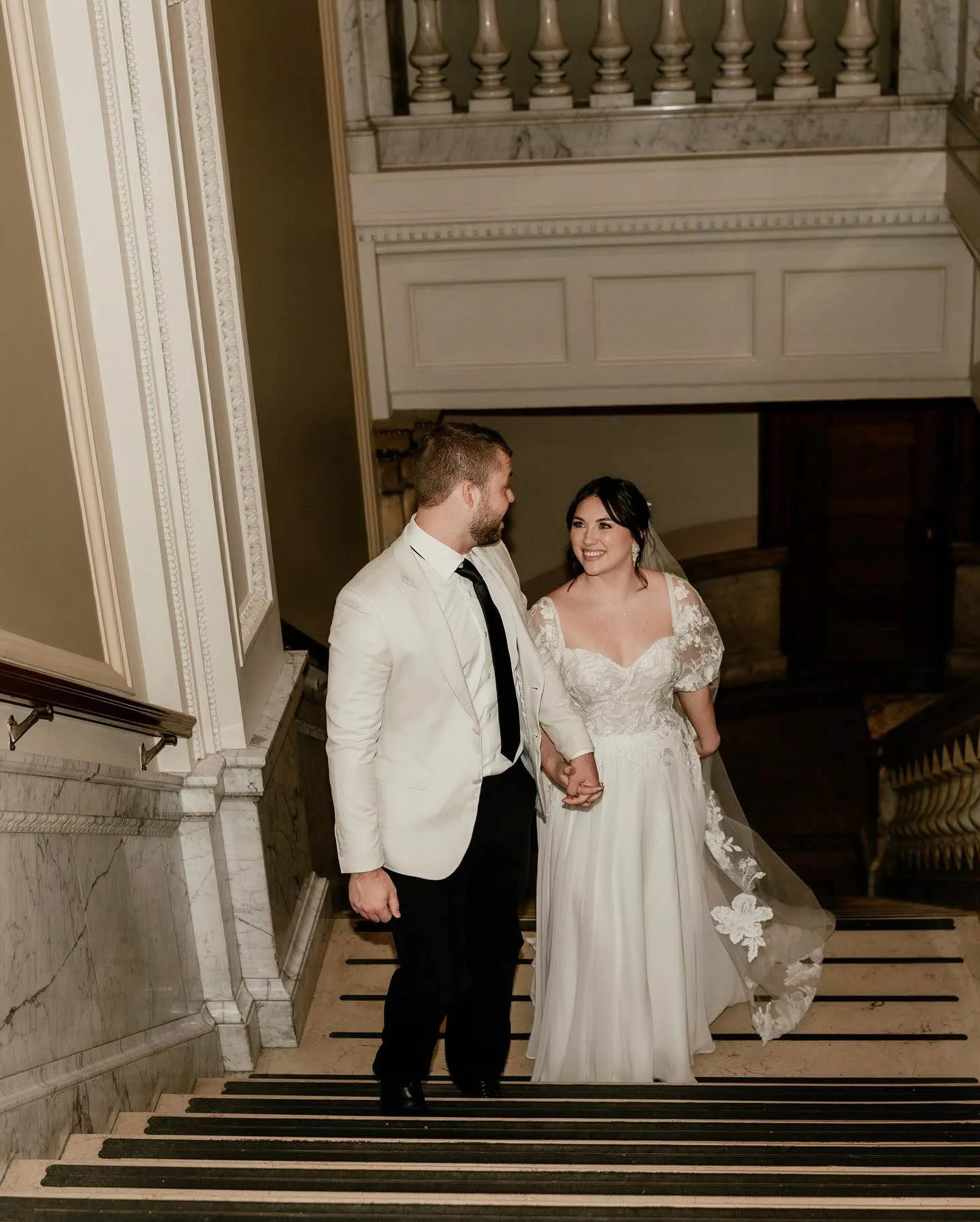MAIE House bridal hair and makeup — bride in lace cap-sleeve gown with veil walking down a marble staircase hand-in-hand with groom