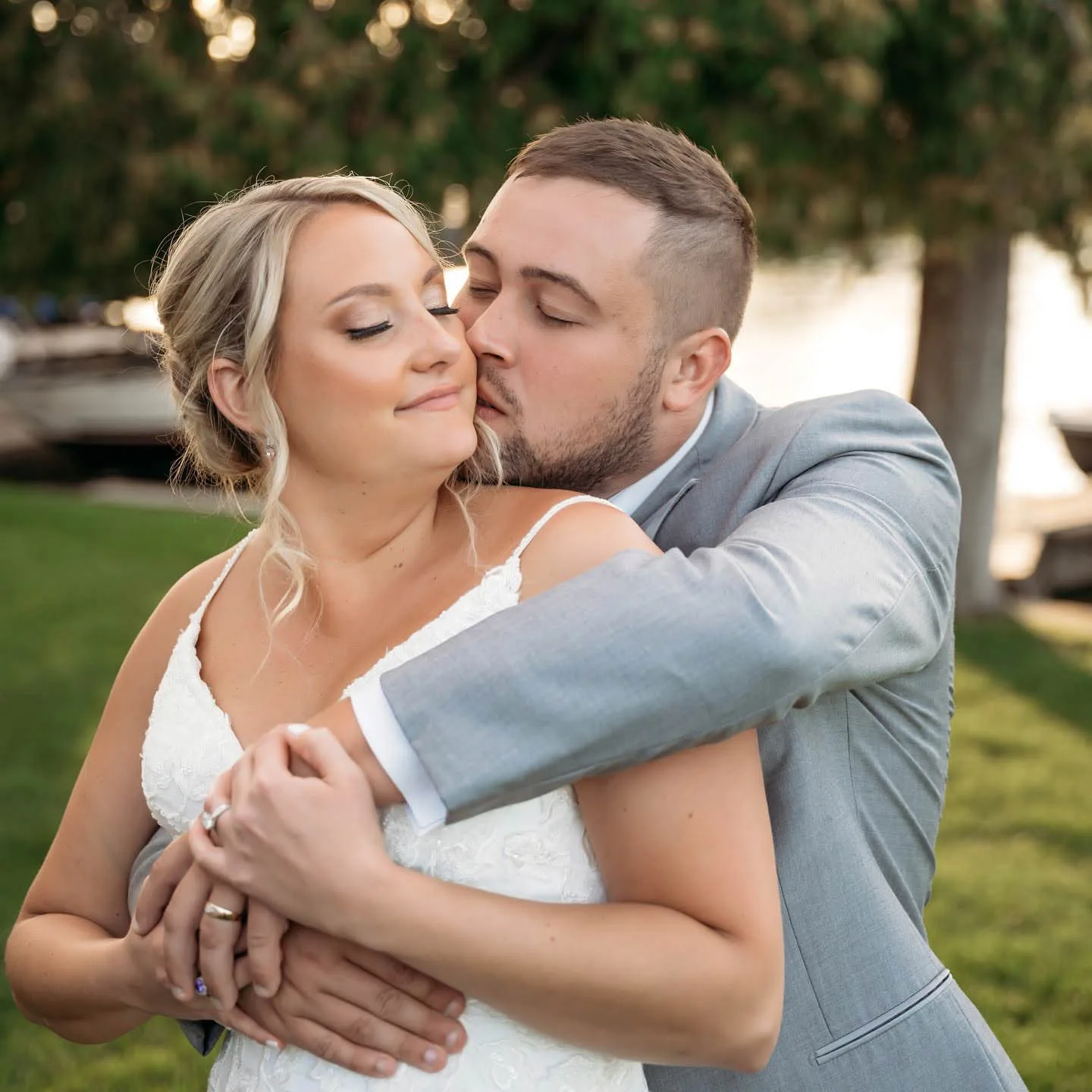 MAIE House soft glam bridal makeup — groom kissing bride's cheek during golden hour portrait by the water