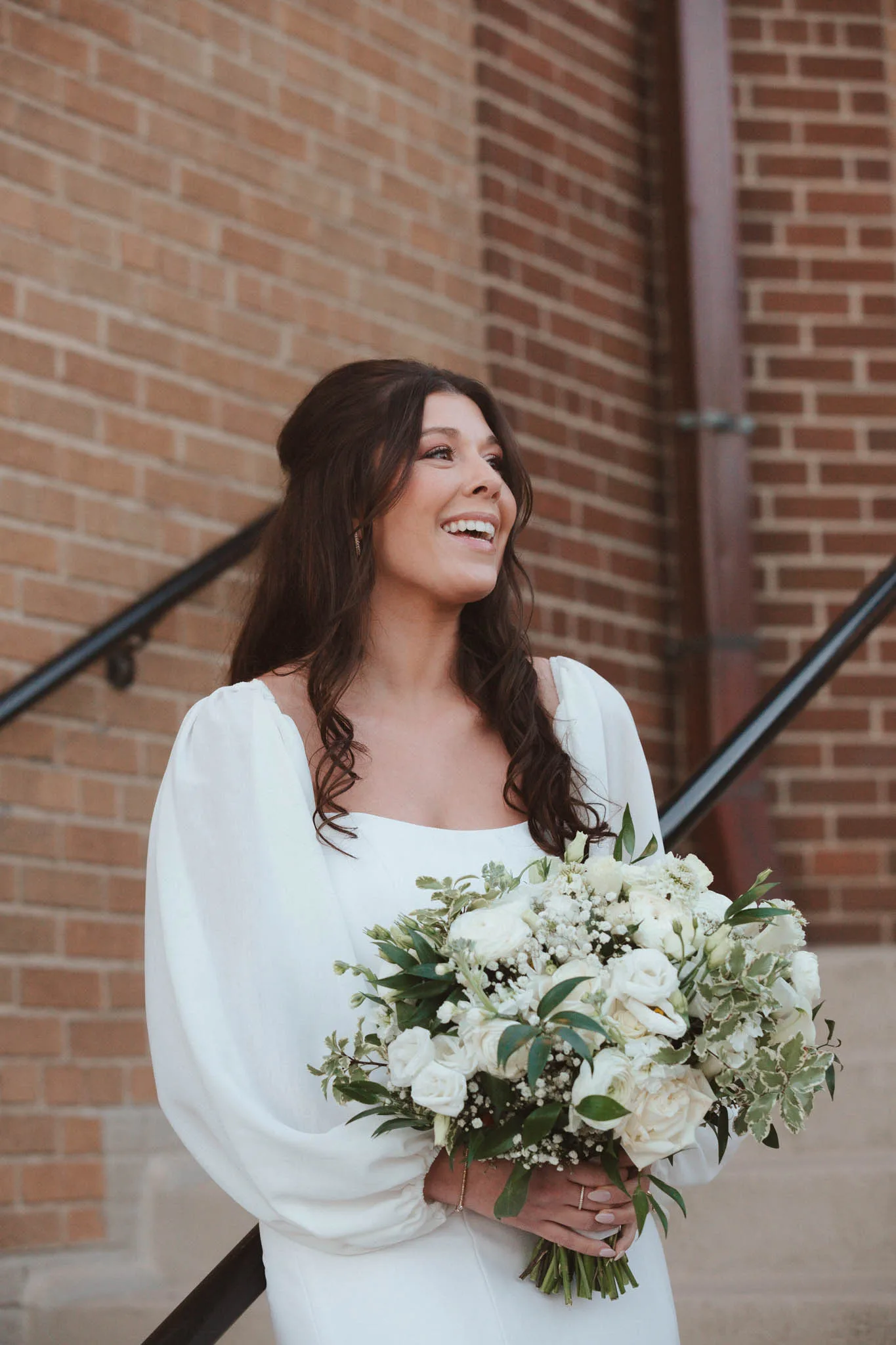 MAIE House bridal hair and natural glam makeup — bride laughing on outdoor steps holding a white and greenery bouquet