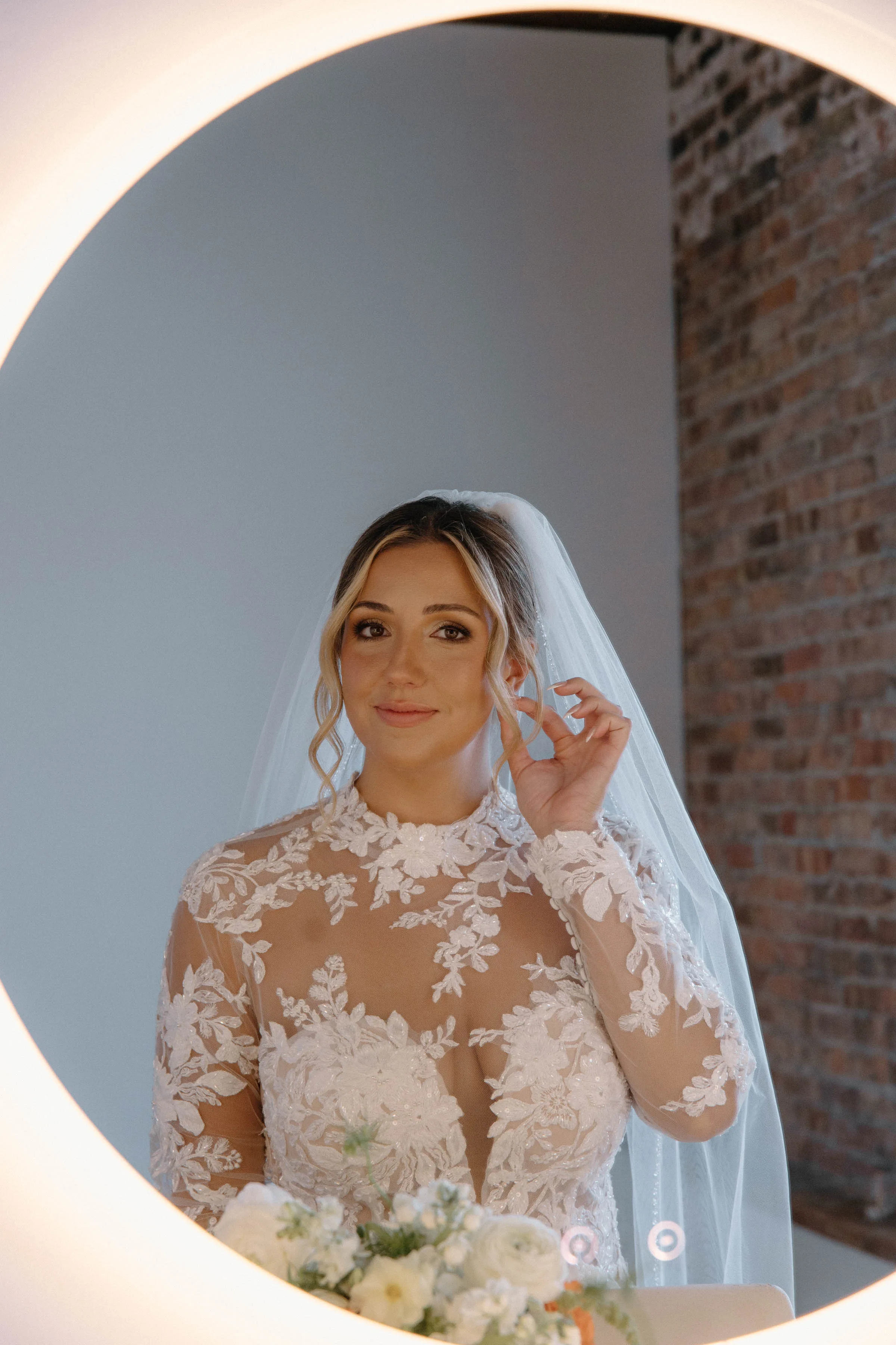 MAIE House bridal makeup and veil styling — closeup portrait of bride in beaded lace gown reflected in a round mirror