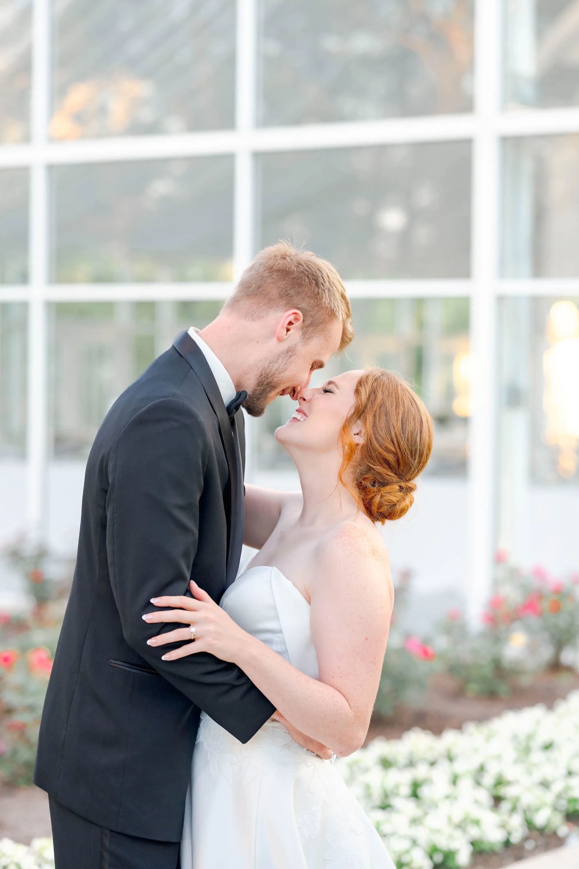 MAIE House bridal updo — bride and groom nose-to-nose laughing together in front of a glass greenhouse with flower garden