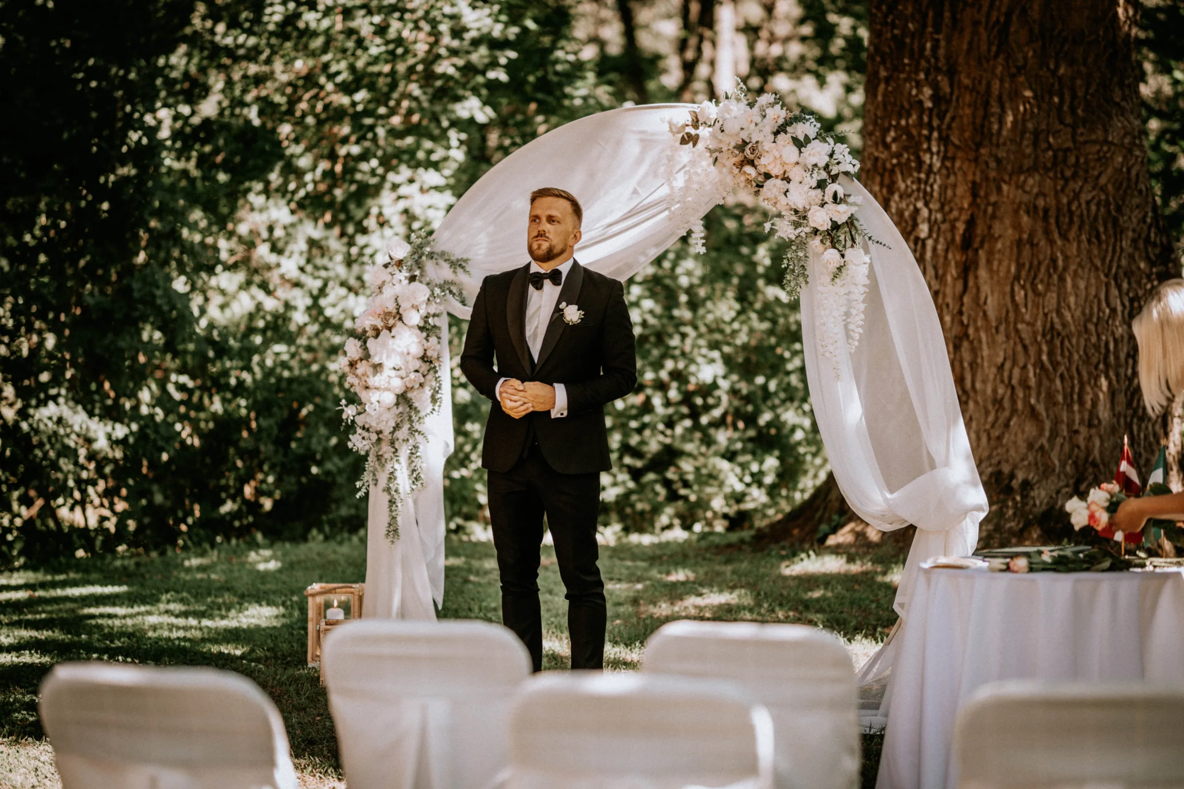 A groom in a black tuxedo stands under a floral wedding arch in