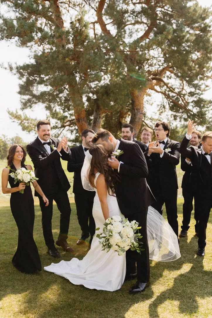 A newlywed couple kisses outdoors while their bridal party, wearing classic black tuxedos and black bridesmaid dresses, cheers and applauds under a large pine tree at a wedding planned by Events by Vanessa.