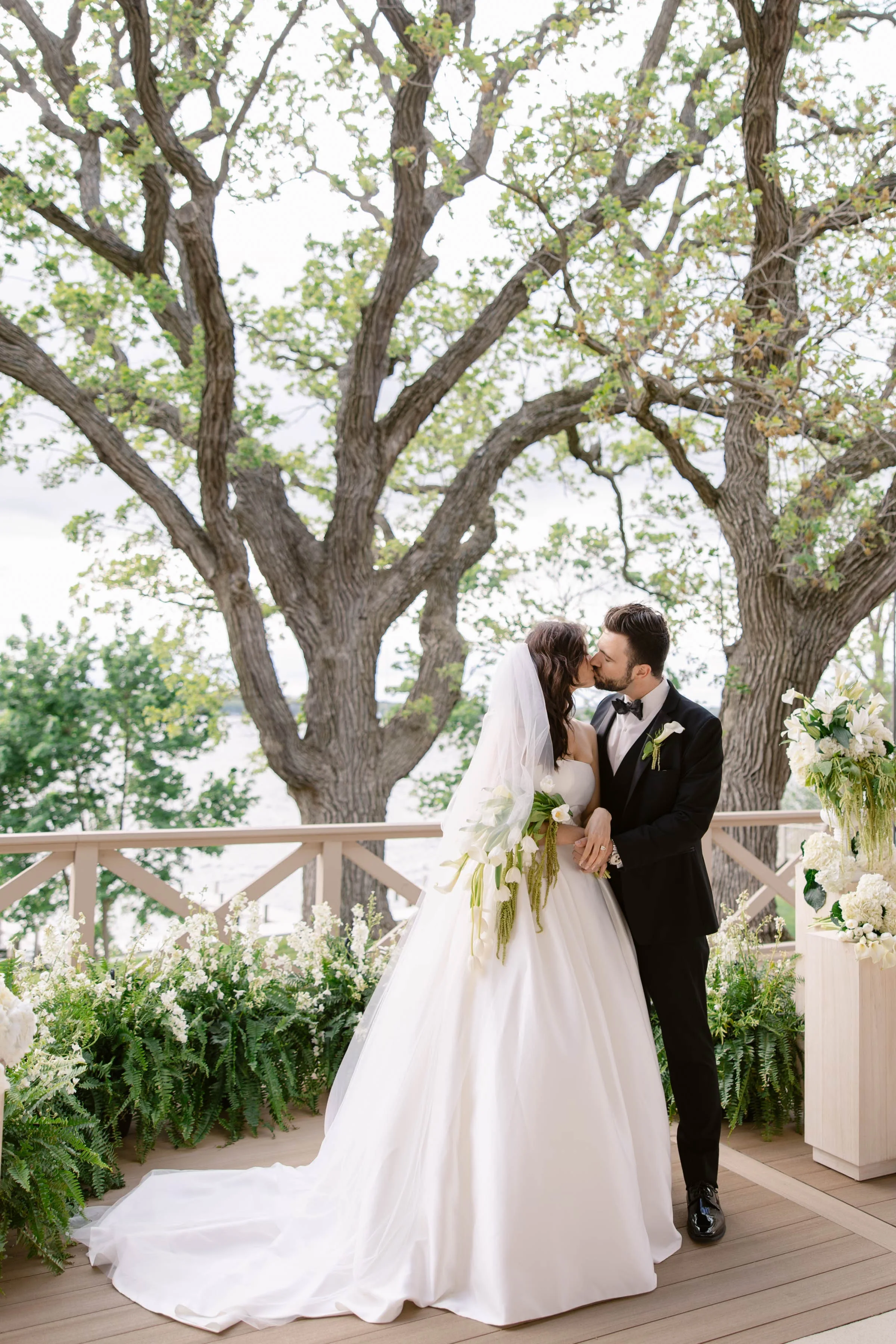 A bride and groom share a kiss on an outdoor wooden deck framed by lush greenery, white floral arrangements, and large oak trees at a Wisconsin wedding designed by Events by Vanessa.