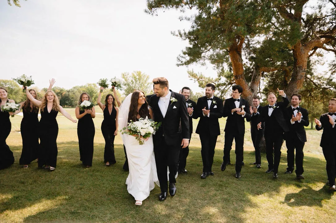 A bride and groom walk hand-in-hand across a green lawn while their wedding party walks behind them celebrating, cheering, and holding white bouquets, coordinated by Events by Vanessa.