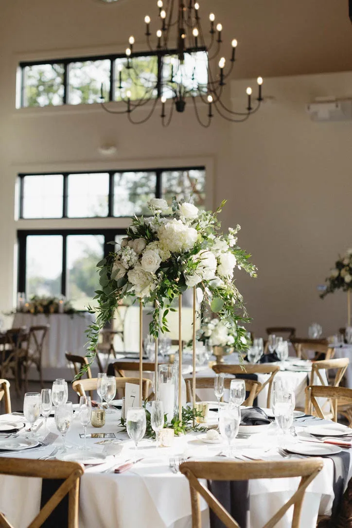 A tall white and green floral centerpiece featuring hydrangeas and roses on a modern gold stand anchors a round reception table with cross-back chairs and a black-and-white runner, styled by Events by Vanessa.