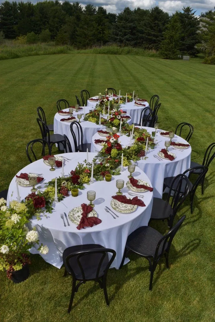 A serpentine wedding reception tablescape on a manicured green lawn, featuring white tablecloths, a continuous runner of greenery with red florals and grapes, burgundy napkins, and black chairs designed by Events by Vanessa.