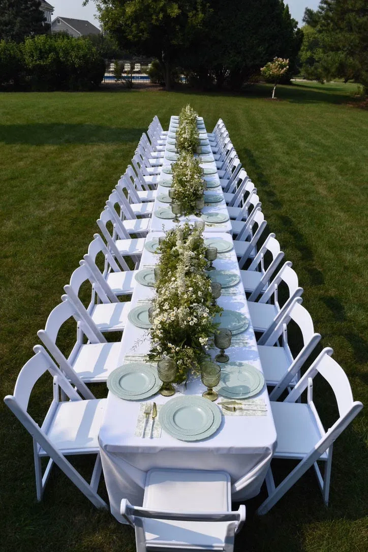 A long banquet-style wedding reception table on a green lawn, styled with white linens, white folding chairs, mint green plates, and a continuous lush runner of white wildflowers and greenery by Events by Vanessa.