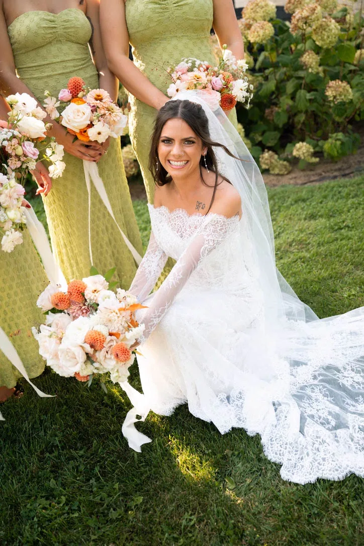 A smiling bride in a lace gown kneels on the grass holding a pastel bouquet with orange dahlias and blush roses, next to bridesmaids in sage green dresses at a wedding styled by Events by Vanessa.