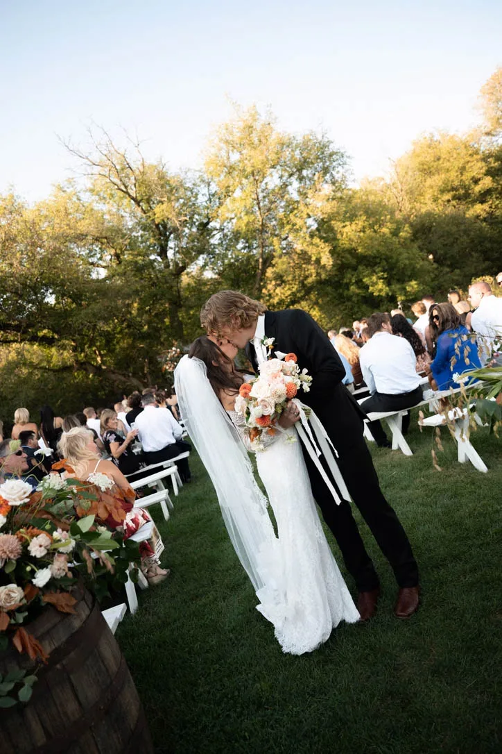 A newlywed couple kisses in the aisle during an outdoor wedding ceremony on a lawn, surrounded by guests on white benches and fall-themed floral barrels, planned by Events by Vanessa.