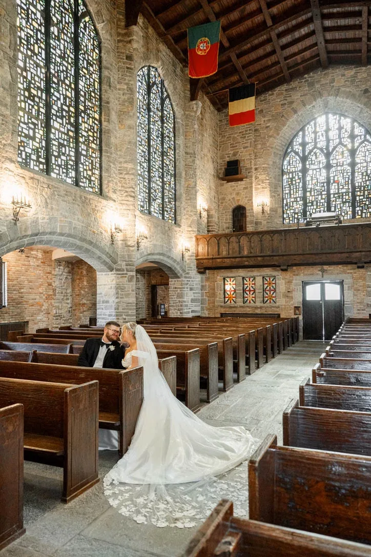 A bride and groom sit together in wooden pews inside a historic stone church featuring vaulted ceilings, towering stained glass windows, and hanging flags, coordinated by Events by Vanessa.