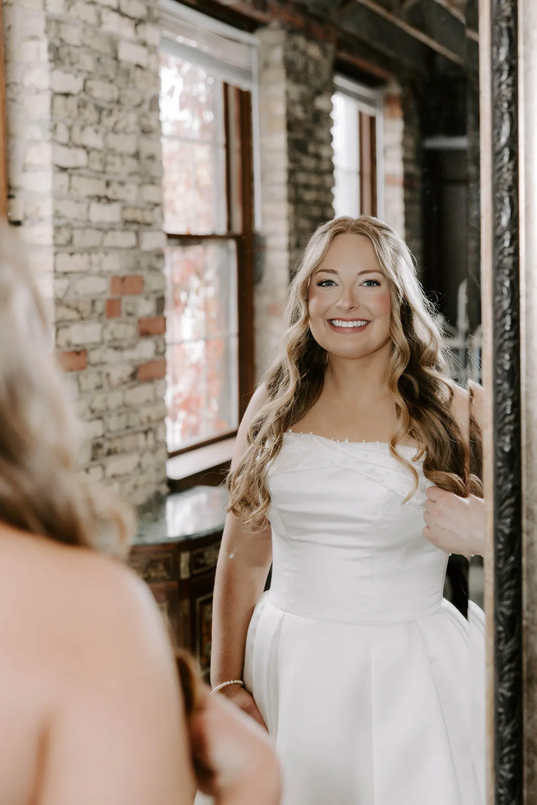 Smiling bride looking into a mirror showing her long wavy bridal hair and soft glam wedding makeup by JJ Bridal Artistry.