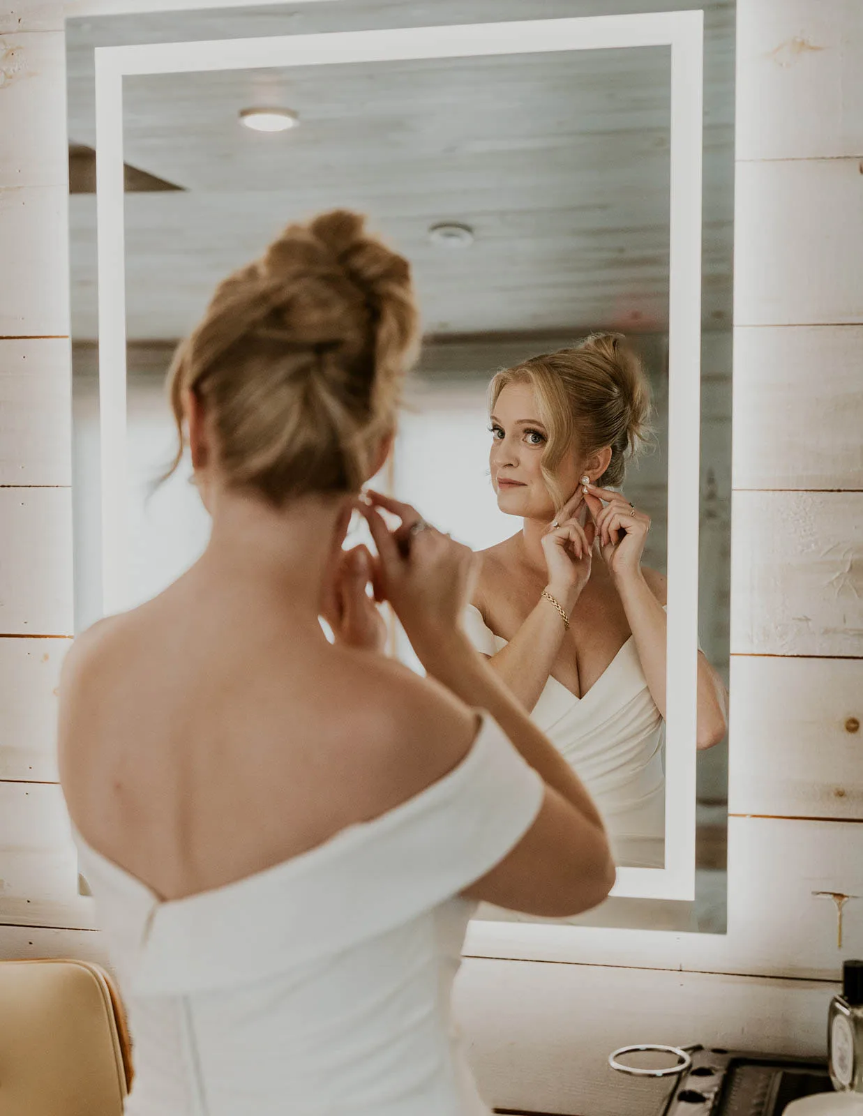 Bride looking in an illuminated mirror putting on earrings with an elegant high bun wedding updo by JJ Bridal Artistry.