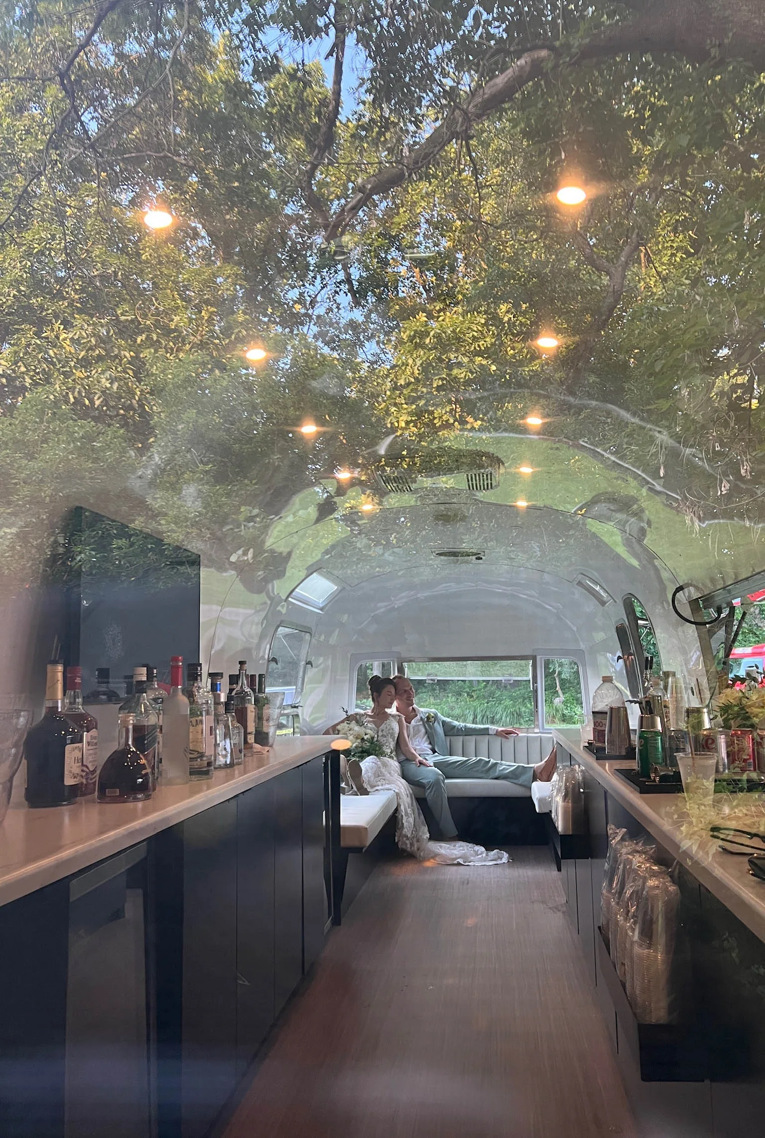 An interior shot of The Airbar taken through the window, capturing a bride and groom relaxing on the built-in lounge seating at the back of the vintage Airstream trailer. The long white countertops in the foreground are stocked with an array of liquor bottles, mixers, and glassware, while the glossy white interior ceiling beautifully reflects the dense green tree canopy and overhead lights outside.