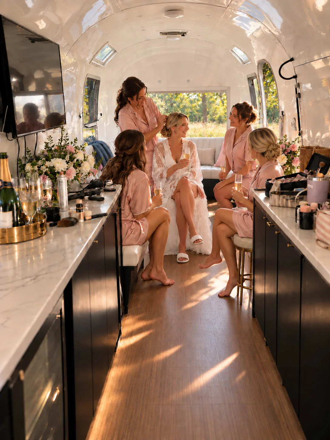 A bright, airy photograph of a bride and her bridesmaids getting ready inside the white, luxury lounge interior of The Airbar. The bride sits in a lace robe holding a glass of champagne while a bridesmaid adjusts her hair, surrounded by four friends in matching pink silk pajamas who are sitting on white leather benches, laughing and talking. The long counters are styled with floral arrangements, champagne glasses, and hair and makeup styling tools.